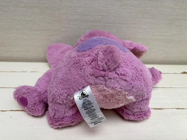 May include: A plush toy in shades of purple, with a darker purple patch on its back. The toy has a soft, furry texture and a Disney tag attached. The toy is lying on a light-colored wooden surface.