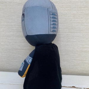 May include: A plush toy featuring a character with a gray helmet-like head, black cape, and gray and yellow accents. The toy is designed with a cartoonish aesthetic and stands upright.