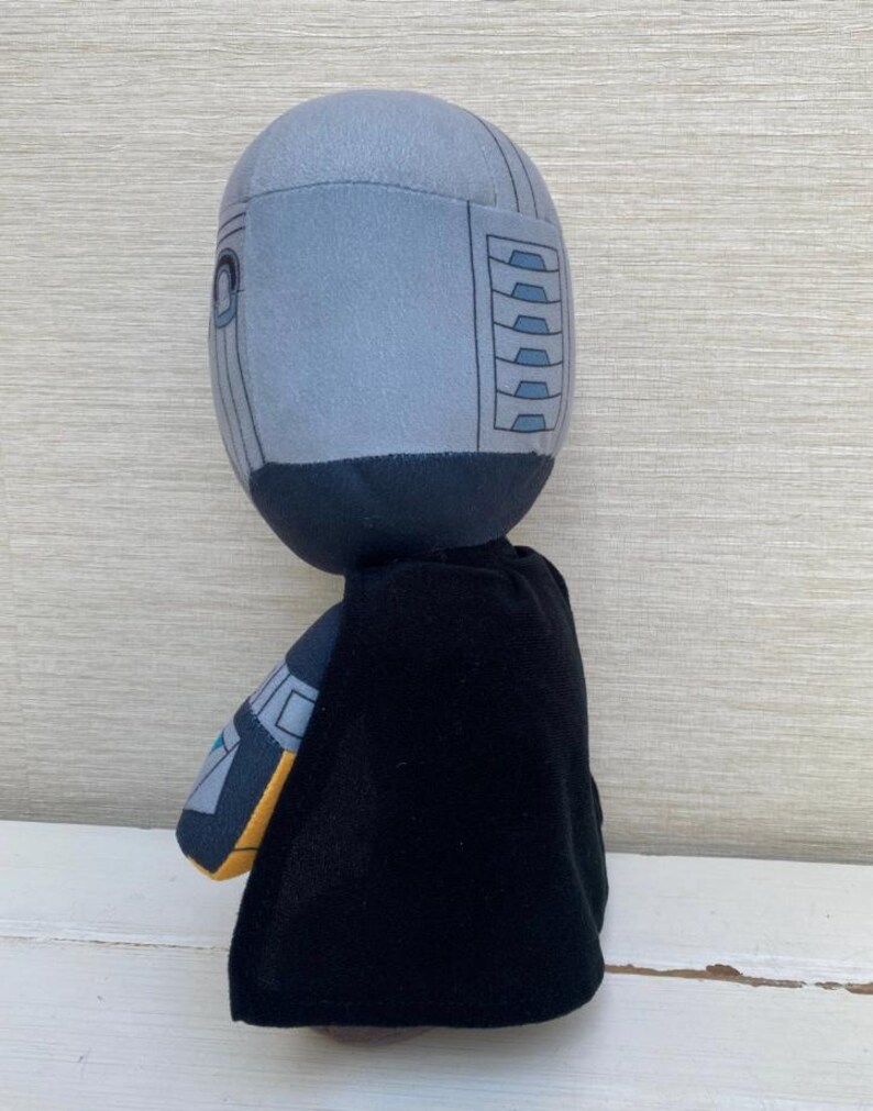 May include: A plush toy featuring a character with a gray helmet-like head, black cape, and gray and yellow accents. The toy is designed with a cartoonish aesthetic and stands upright.