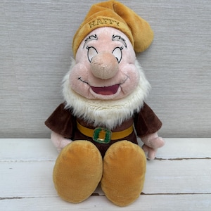 Puede incluir: Muñeco de peluche del personaje de Disney Happy, uno de los Siete Enanitos. El muñeco tiene un gorro amarillo con "HAPPY" bordado, una barba blanca y un atuendo marrón con un cinturón dorado. La cara del muñeco es rosa con una nariz grande y una expresión sonriente.
