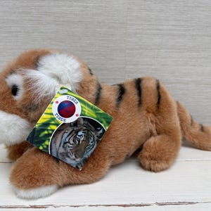 Op de afbeelding: Een pluche tijger met bruin en wit haar, zwarte strepen en een label met een tijgergezicht. Het speelgoed ligt op een wit houten oppervlak.