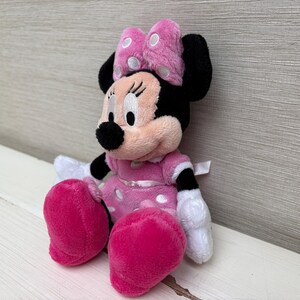 May include: A plush Minnie Mouse doll, featuring a pink dress with white polka dots, matching pink bow with white polka dots, and large pink shoes. The doll has black ears, a black nose, and white gloves. The doll is sitting on a white surface.