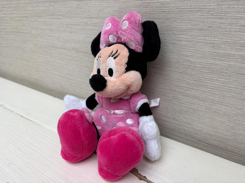 May include: A plush Minnie Mouse doll, featuring a pink dress with white polka dots, matching pink bow with white polka dots, and large pink shoes. The doll has black ears, a black nose, and white gloves. The doll is sitting on a white surface.