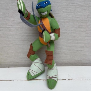 May include: A plush Leonardo Teenage Mutant Ninja Turtle doll. The doll is green with an orange chest, brown accents, and a blue mask. It is holding two grey sai weapons and a brown staff. The doll is standing on a white surface.