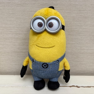 May include: A yellow plush toy character with a cylindrical body, wearing blue denim-textured overalls with a central logo and black gloves and shoes. The figure features large, round, silver-rimmed goggles over brown eyes and a small smile.