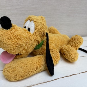 Pode incluir: Um peluche de cão com pelo amarelo dourado, orelhas pretas compridas, nariz preto e olhos brancos. A personagem está representada numa posição deitada com a língua cor-de-rosa de fora e uma coleira verde à volta do pescoço.