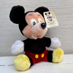 Puede incluir: Un peluche de Mickey Mouse con orejas negras, cuerpo negro, pantalones cortos rojos con botones amarillos y pies amarillos. El juguete tiene una etiqueta con una ilustraci&oacute;n de Mickey Mouse. El juguete est&aacute; sobre una superficie blanca.