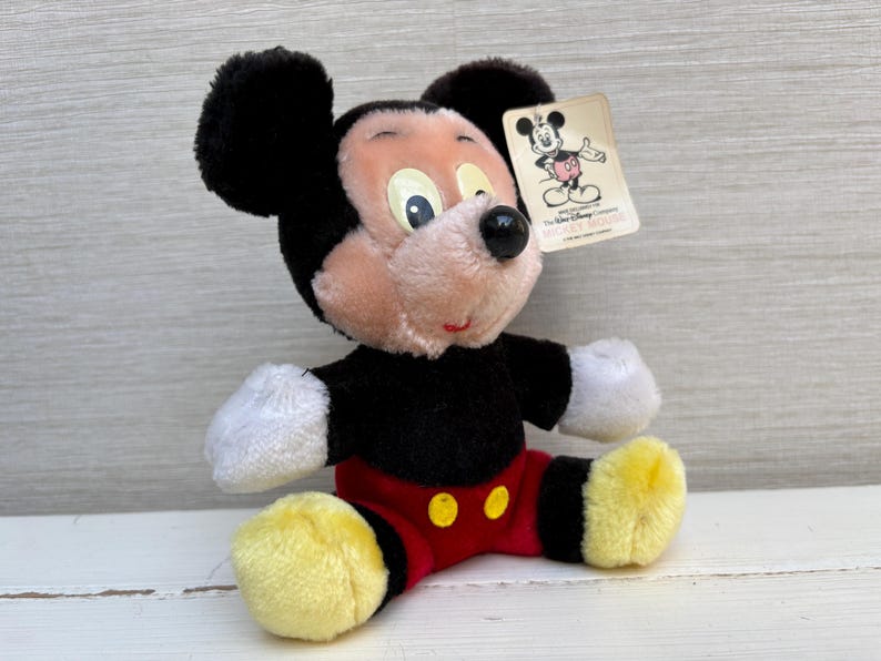 Puede incluir: Un peluche de Mickey Mouse con orejas negras, cuerpo negro, pantalones cortos rojos con botones amarillos y pies amarillos. El juguete tiene una etiqueta con una ilustraci&oacute;n de Mickey Mouse. El juguete est&aacute; sobre una superficie blanca.