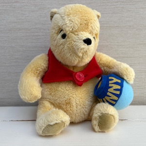 Puede incluir: Un oso de peluche con pelaje amarillo claro que lleva un chaleco rojo con un botón rojo. El oso sostiene una maceta azul con un borde amarillo y el texto "HUNNY" impreso en letras amarillas en el lateral. El oso tiene ojos y nariz negros.