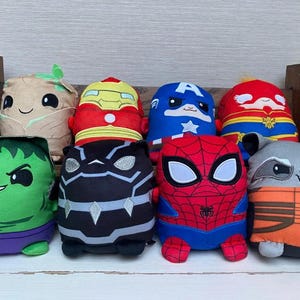 May include: A collection of eight plush toys featuring various Marvel characters. The toys are colorful, with designs including Hulk, Black Panther, Spider-Man, Rocket, Captain America, Iron Man, Captain Marvel, and Groot. They are arranged on a white surface.