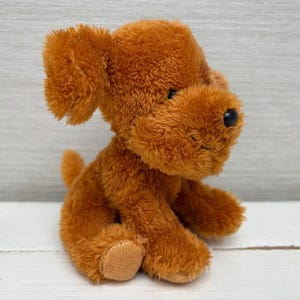 May include: A soft, brown plush dog toy. The toy dog has floppy ears, a small tail, and a black nose. The dog is sitting on a white surface. The toy has a soft, textured appearance.