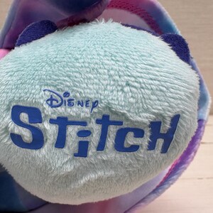 May include: Close-up of a plush toy featuring the Disney Stitch logo in blue on a light blue, textured surface. The toy has blue ears and is surrounded by colorful fabric in shades of pink, purple, and blue.