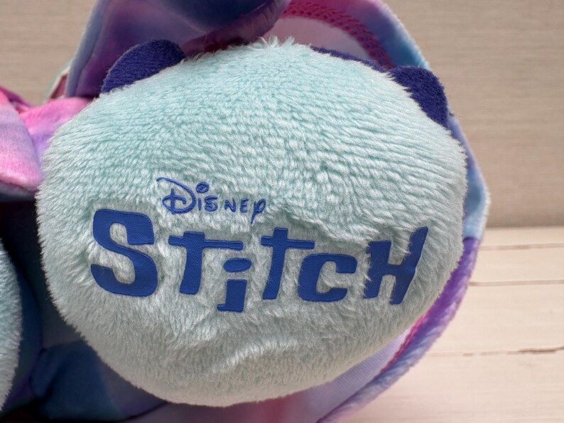 May include: Close-up of a plush toy featuring the Disney Stitch logo in blue on a light blue, textured surface. The toy has blue ears and is surrounded by colorful fabric in shades of pink, purple, and blue.