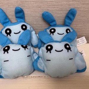 May include: Four plush toys shaped like blue bunnies with large ears and smiling faces. The bunnies have black eyes and eyebrows, and a light blue body. The toys are made of soft fabric and are suitable for children.