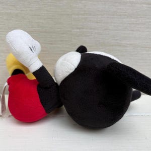 Op de afbeelding: Een pluche speelgoed van een stripfiguur, met een zwart lichaam, witte handschoenen en een rode onderkant. Het speelgoed ligt op zijn zij, met zijn witte handschoenhand omhoog. Het speelgoed heeft een gele kop en een wit gezicht.