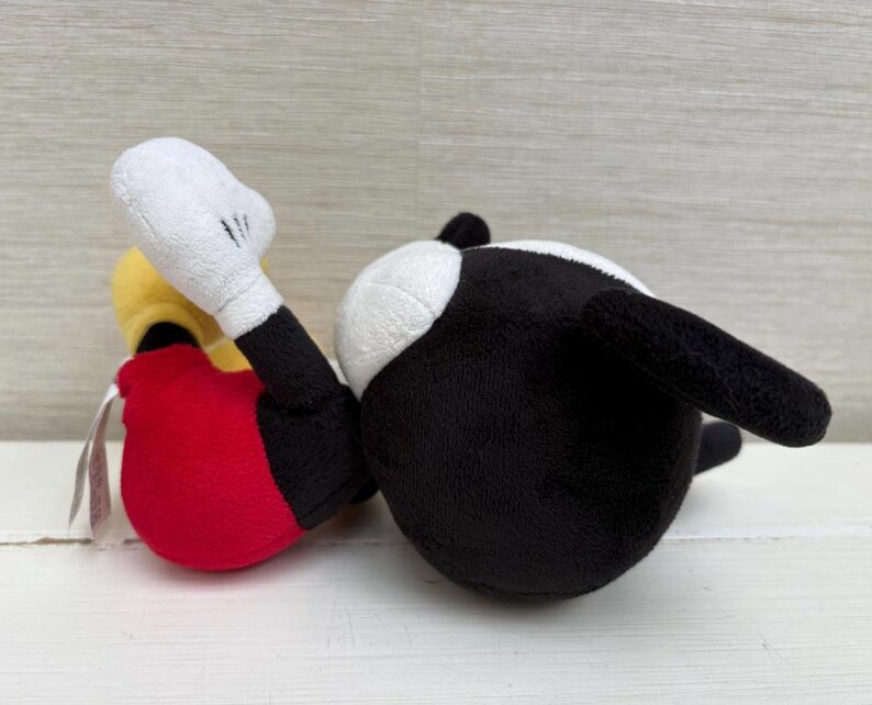 Op de afbeelding: Een pluche speelgoed van een stripfiguur, met een zwart lichaam, witte handschoenen en een rode onderkant. Het speelgoed ligt op zijn zij, met zijn witte handschoenhand omhoog. Het speelgoed heeft een gele kop en een wit gezicht.