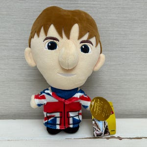 May include: A plush doll featuring a character with brown hair, large dark eyes, and a prominent nose. The doll wears a jacket printed with the Union Jack flag in red, white, and blue, and includes a gold circular tag attached to the side.