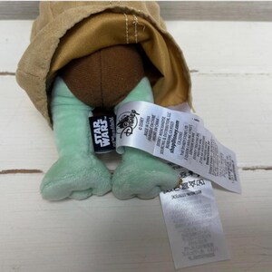 May include: A plush toy featuring the character from the Star Wars series. The toy has light green feet, a brown head, and a tan-colored robe. The Star Wars and Disney logos are visible on a tag attached to the toy.