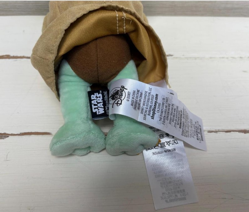 May include: A plush toy featuring the character from the Star Wars series. The toy has light green feet, a brown head, and a tan-colored robe. The Star Wars and Disney logos are visible on a tag attached to the toy.