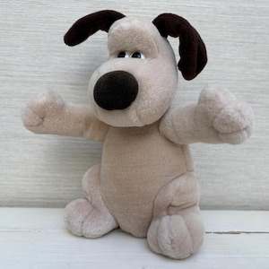 May include: A plush dog toy featuring a light beige body, dark brown floppy ears, and a large, rounded dark brown nose. The stuffed animal has small black eyes, a soft fabric texture, and is posed with its arms extended outward to the sides.