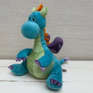 May include: A plush dragon toy featuring a bright blue body with a light green textured belly. It has small purple wings, purple claws on its feet and hands, and a row of orange spikes running down its back. The dragon has a rounded head.