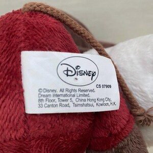 May include: A close-up of a plush toy in red, brown, and white. A white tag with the Disney logo and text is attached. The toy has a braided brown cord.