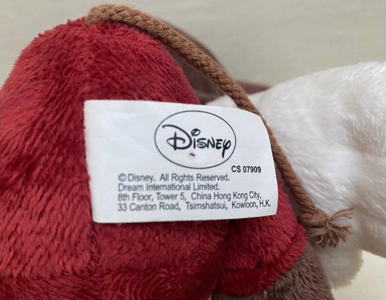 May include: A close-up of a plush toy in red, brown, and white. A white tag with the Disney logo and text is attached. The toy has a braided brown cord.