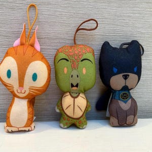Puede incluir: Tres peluches: un gato naranja con orejas rosas, una tortuga verde con caparazón marrón y un perro negro con máscara y capa azules. Cada juguete tiene un lazo para colgar.