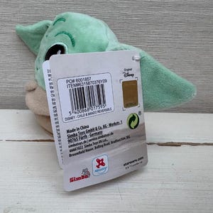 May include: A green and tan plush toy of a character from The Mandalorian series, with a tag attached. The tag includes text and a barcode. The toy has large ears.
