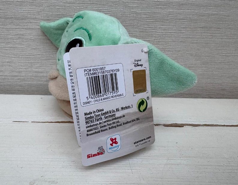 May include: A green and tan plush toy of a character from The Mandalorian series, with a tag attached. The tag includes text and a barcode. The toy has large ears.