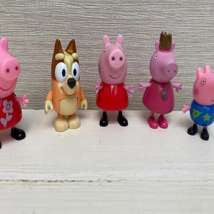 May include: Five colourful figurines of cartoon characters. The characters include two pink pigs in red dresses, a dog, a pig wearing a crown, and a pig in a blue shirt. The figurines are standing on a light-coloured surface.
