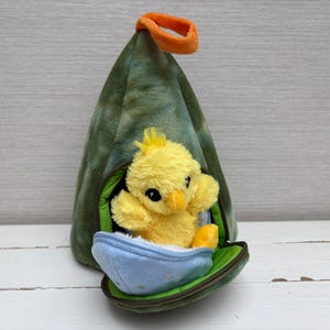 May include: A soft yellow chick toy is nestled inside a green and blue zippered pouch. The pouch features an orange handle and a blue egg-shaped base. The chick has black eyes and a yellow beak.