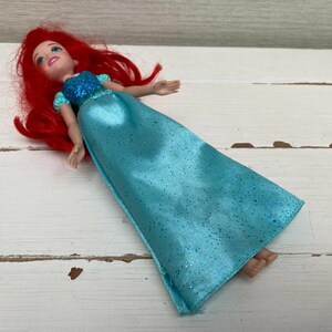 Op de afbeelding: Pop van een personage met rood haar, gekleed in een blauwe jurk met glitteraccenten. De pop heeft een glimlachende uitdrukking en ligt op een wit houten oppervlak.