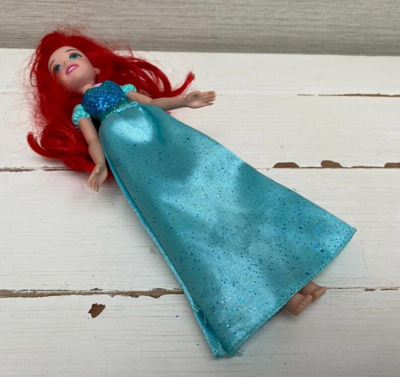 Op de afbeelding: Pop van een personage met rood haar, gekleed in een blauwe jurk met glitteraccenten. De pop heeft een glimlachende uitdrukking en ligt op een wit houten oppervlak.