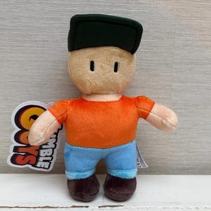 May include: A plush toy character with a tan face, orange shirt, blue pants, and a dark green cap. The toy has small black eyes and brown shoes. A tag with the words "Stumble Guys" is attached.