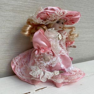 May include: A vintage porcelain doll in a pink dress and matching hat, trimmed with white lace. The doll has blonde curly hair and is wearing a satin dress with lace accents. A decorative collectible item.