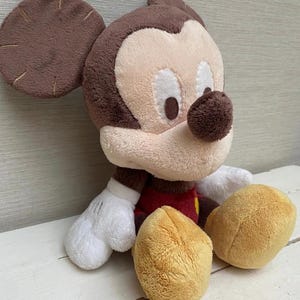 May include: A soft toy of Mickey Mouse, featuring brown ears, a tan face, and a black nose. He is dressed in a red shirt with yellow buttons, white gloves, and tan shorts. The toy is placed on a white surface.