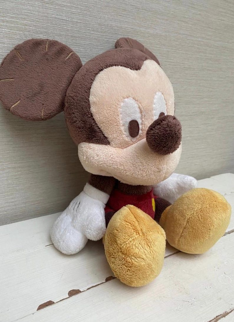 May include: A soft toy of Mickey Mouse, featuring brown ears, a tan face, and a black nose. He is dressed in a red shirt with yellow buttons, white gloves, and tan shorts. The toy is placed on a white surface.