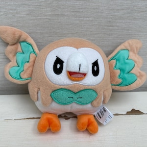May include: A plush toy shaped like a round owl with tan fur, a white face, and large black eyes. It features mint green wing accents and a matching bow-tie shape on its chest. The toy has bright orange feet and a small white tag attached to the side.