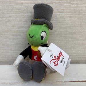 May include: A plush Jiminy Cricket toy featuring a green head, a grey top hat, a black jacket with a red vest, and white gloves. The character has large eyes and is sitting down. A white tag attached to the arm displays the text The Disney Store.