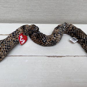 Op de afbeelding: Een pluche slangenspeelgoed met een bruin en zwart patroon. De slang heeft oranje ogen, een rode mond en een rood hartvormig label. Het speelgoed is gemaakt van zacht materiaal en is ontworpen om op een slang te lijken.