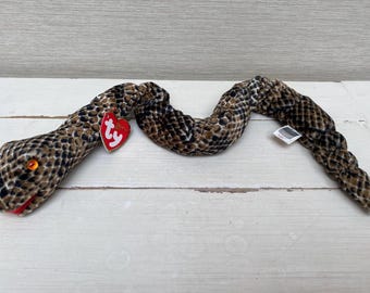 Pode incluir: Um brinquedo de pelúcia em forma de cobra com um padrão marrom e preto. A cobra tem olhos laranja, boca vermelha e uma etiqueta vermelha em forma de coração. O brinquedo é feito de material macio e foi projetado para se assemelhar a uma cobra.