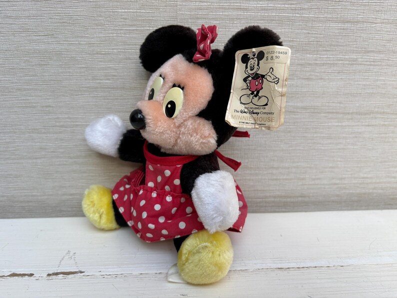 K&ouml;nnte beinhalten: Ein Vintage-Minnie-Mouse-Pl&uuml;schtier. Das Pl&uuml;schtier hat ein rotes Kleid mit wei&szlig;en Punkten, gelbe Schuhe und eine rote Schleife. Das Spielzeug hat ein Etikett mit einer Mickey-Mouse-Illustration und dem Text "MINNIE MOUSE".