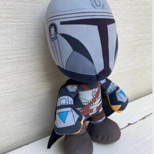 May include: A plush toy depicting a character from a popular science fiction series. The figure has a gray helmet, black and gray armor, and brown boots. The toy is standing on a white surface.