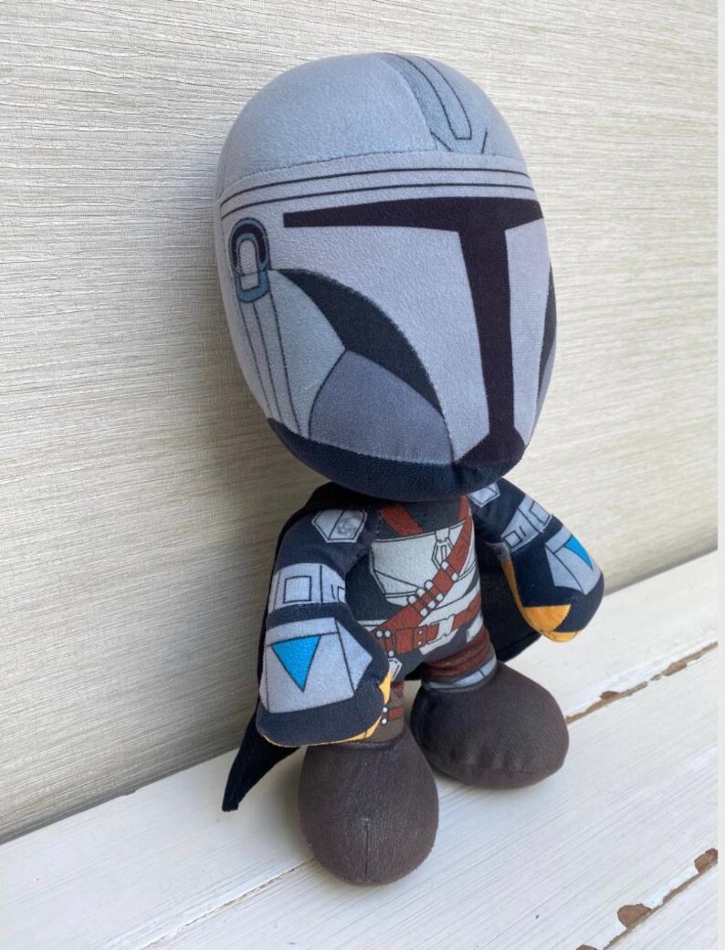 May include: A plush toy depicting a character from a popular science fiction series. The figure has a gray helmet, black and gray armor, and brown boots. The toy is standing on a white surface.