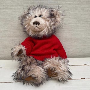 May include: A plush teddy bear with a shaggy, two-tone fur coat in shades of gray and cream. The bear wears a bright red sweater. The bear's paws and snout are a light brown color. The toy is sitting on a white surface.