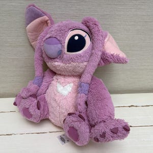 May include: A plush toy of the character Angel from the Disney movie Lilo & Stitch. The toy is primarily purple and pink, with large black eyes, long ears, and a heart-shaped design on its chest. The plush is soft and cuddly.