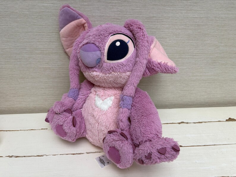 May include: A plush toy of the character Angel from the Disney movie Lilo & Stitch. The toy is primarily purple and pink, with large black eyes, long ears, and a heart-shaped design on its chest. The plush is soft and cuddly.