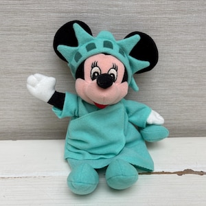 Może przedstawiać: Pluszowa lalka Minnie Mouse przebrana za Statuę Wolności. Lalka ma r&oacute;żową twarz, czarne uszy i nosi turkusową szatę i koronę. Jej prawa ręka jest uniesiona, a lewa ręka jest po jej stronie.