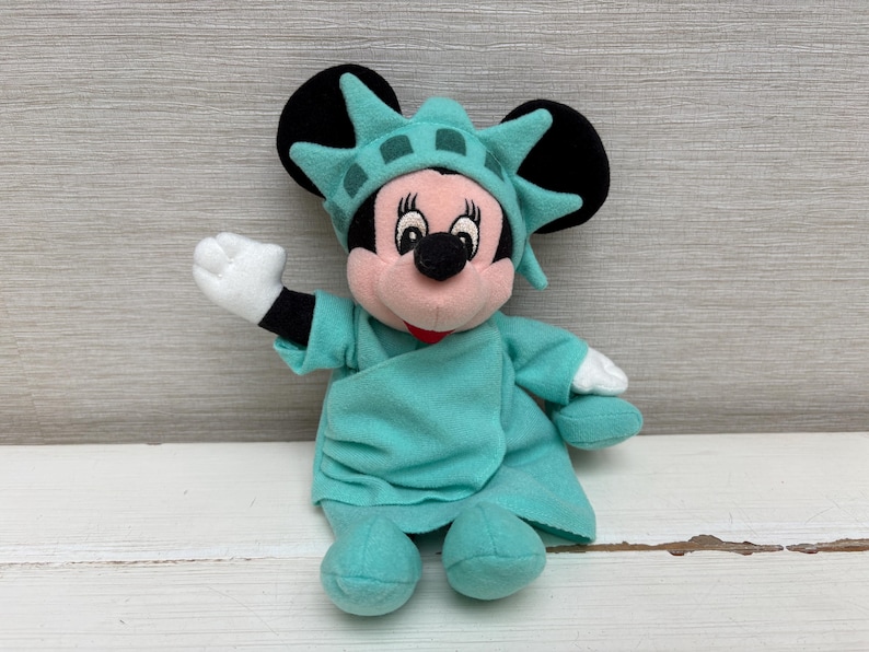 Może przedstawiać: Pluszowa lalka Minnie Mouse przebrana za Statuę Wolności. Lalka ma r&oacute;żową twarz, czarne uszy i nosi turkusową szatę i koronę. Jej prawa ręka jest uniesiona, a lewa ręka jest po jej stronie.